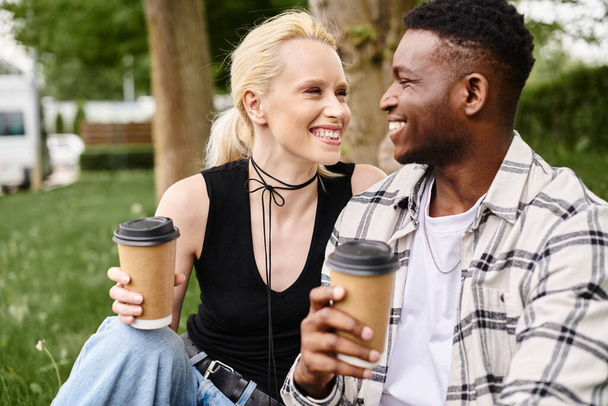 Çok kültürlü bir çift, Afro-Amerikalı bir erkek ve Kafkasyalı bir kadın, parktaki yemyeşil çimlerin üzerinde oturmuş kahvelerini yudumluyorlar.. - Fotoğraf, Görsel