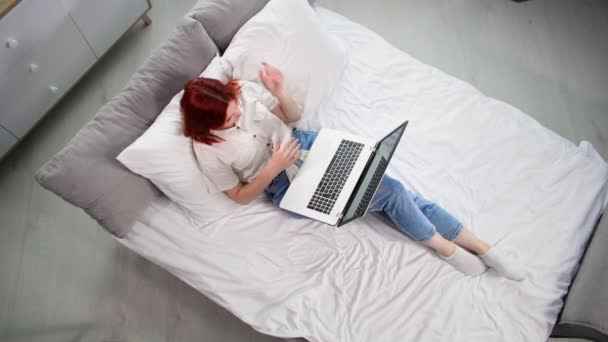 чарівна молода жінка весело спілкується на ноутбуці з друзями, лежачи на ліжку в кімнаті, вид зверху - Кадри, відео