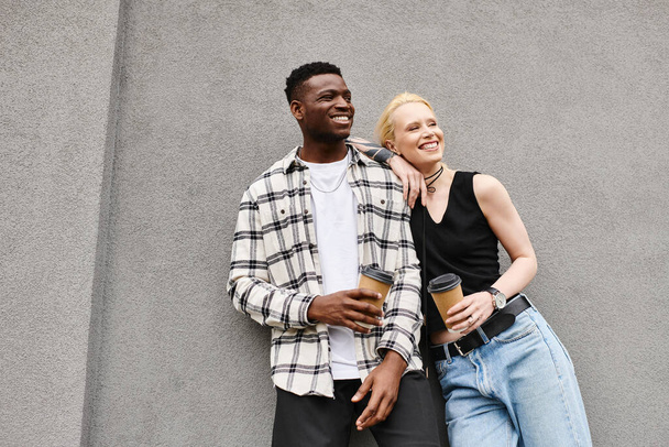 Ένα πολυπολιτισμικό ζευγάρι, ένας άντρας και μια γυναίκα, να στέκονται ευτυχισμένοι μαζί σε έναν αστικό δρόμο δίπλα σε ένα γκρίζο κτίριο.. - Φωτογραφία, εικόνα