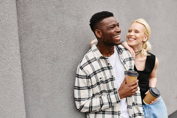 Ένα πολυπολιτισμικό ζευγάρι, ένας άντρας και μια γυναίκα, στέκονται ευτυχισμένοι μαζί σε έναν αστικό δρόμο δίπλα σε ένα γκρίζο κτίριο.. - Φωτογραφία, εικόνα
