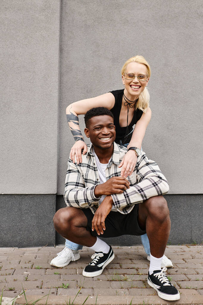 Un alegre novio multicultural y novia posan juntos en una calle urbana cerca de un edificio gris, sonriendo a la cámara. - Foto, Imagen