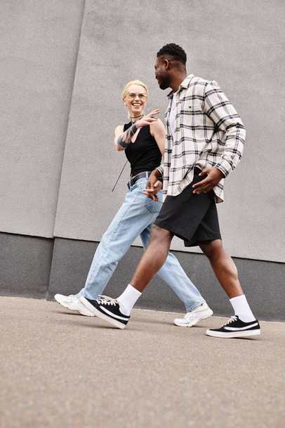 Una coppia felice e multiculturale cammina lungo una strada urbana accanto a un edificio grigio, godendosi reciprocamente la compagnia. - Foto, immagini