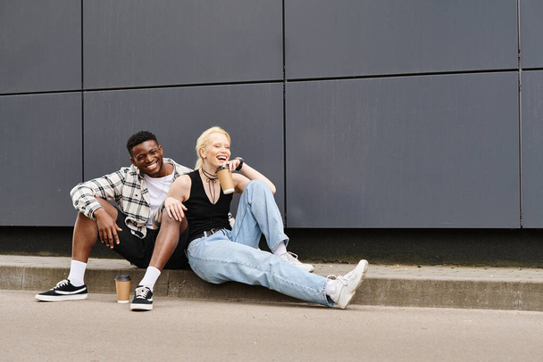 A happy multicultural couple sitting beside each other on the ground near a grey urban building - Fotografie, Obrázek