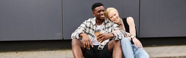 Счастливая, мультикультурная пара наслаждается веселым моментом вместе, сидя рядом с серым зданием. - Фото, изображение