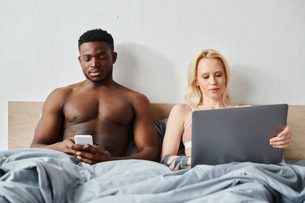 A multicultural boyfriend and girlfriend sit on a bed, focused on their devices. - Foto, afbeelding