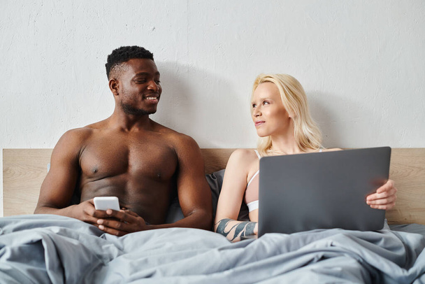 Wielokulturowy mężczyzna i kobieta siedzą blisko na łóżku, pochłonięci ekranem laptopa przed nimi. - Zdjęcie, obraz
