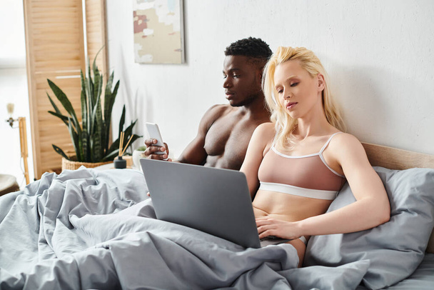 A multicultural man and woman are sitting on a bed, engrossed in the screen of a laptop in an intimate and cozy setting. - Foto, afbeelding
