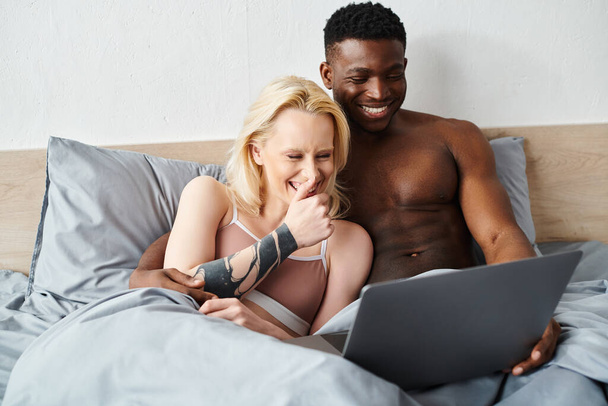 Wielokulturowy mężczyzna i kobieta leżący w łóżku, pochłonięci ekranem laptopa. - Zdjęcie, obraz
