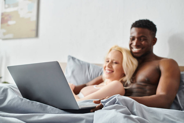 Ein sexy multikulturelles Paar sitzt eng auf einem Bett und ist fasziniert von den Inhalten auf einem Laptop-Bildschirm vor sich.. - Foto, Bild