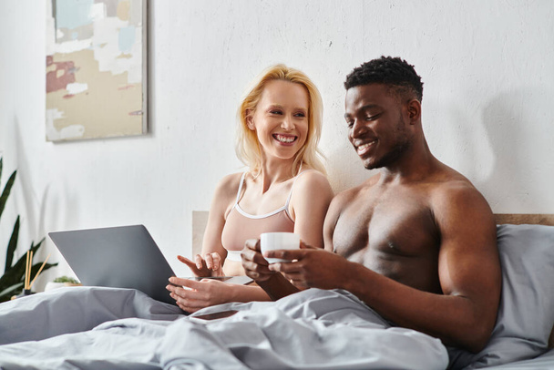 Un fidanzato multiculturale e una fidanzata si siedono su un letto, affascinati dallo schermo di un computer portatile. - Foto, immagini