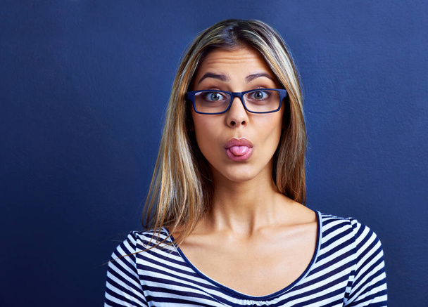 Funny, face and woman with glasses in studio for tongue out, silly or humor on blue background. Need, portrait and female model with emoji expression, gesture or geek, aesthetic or quirky personality. - Photo, Image