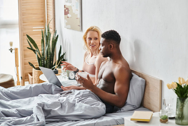 Мультикультурный парень и девушка сидят на кровати, погруженные в экран ноутбука. - Фото, изображение