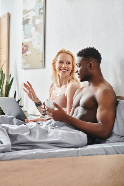 A sexy multicultural couple, boyfriend and girlfriend, sitting on a bed, focused on a laptop screen together. - Foto, afbeelding