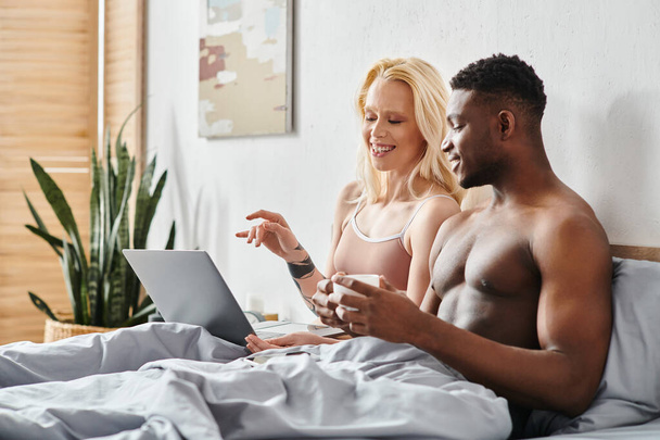 Um homem e uma mulher, um namorado multicultural e uma namorada, confortavelmente sentados em uma cama, intensamente focados em uma tela de laptop. - Foto, Imagem