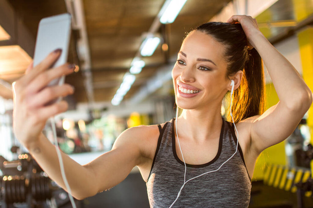 Portrait de belle fille sportive souriante avec écouteur prenant un selfie en utilisant un téléphone intelligent dans la salle de gym. - Photo, image
