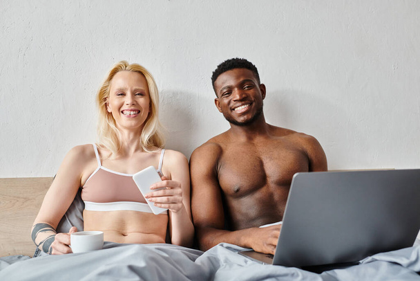 Um casal multicultural sentado em uma cama, absorvido em um laptop. - Foto, Imagem