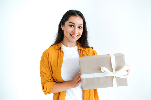 Gifts and surprises concept. Excited happy brunette indian or arabian woman in casual orange shirt, stands on isolated white background, holding big gift box, looking at camera and smiles friendly - Photo, Image