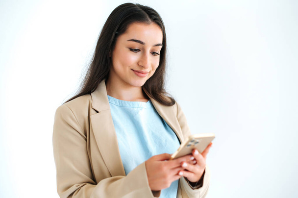 Позитивная довольно индийская или арабская женщина в костюме, используя смартфон для общения с друзьями, просмотра интернета, поиска информации, стоя на изолированном белом фоне, счастливо улыбаясь - Фото, изображение