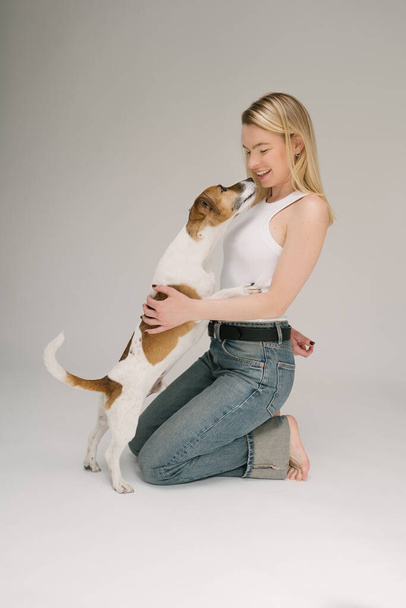 Dog kissing blonde woman pet owner. Happy laughing girl hugging small dog. Love emotions. Studio shot. Grey background - Photo, Image
