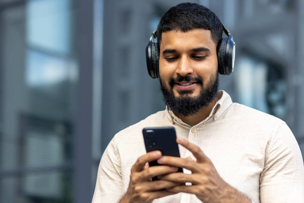 A cheerful young man with a beard is seen outside using his smartphone and listening to music through over-ear headphones, exuding a vibe of relaxation and enjoyment in an urban setting. - Zdjęcie, obraz