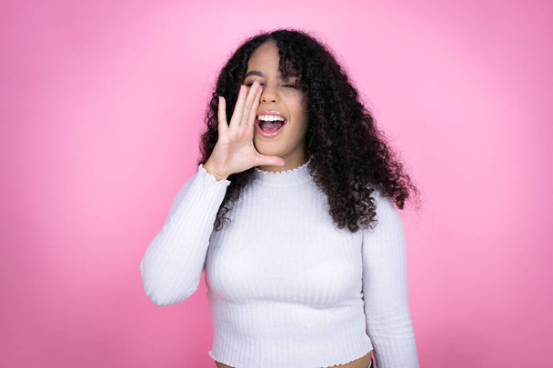 ピンクの背景にカジュアルなセーターを身に着けているアフリカ系アメリカ人女性が叫び声を上げ,口に手で大声で叫ぶ - 写真・画像