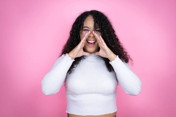 donna afroamericana indossa maglione casual su sfondo rosa urlando e urlando forte a lato con le mani in bocca - Foto, immagini