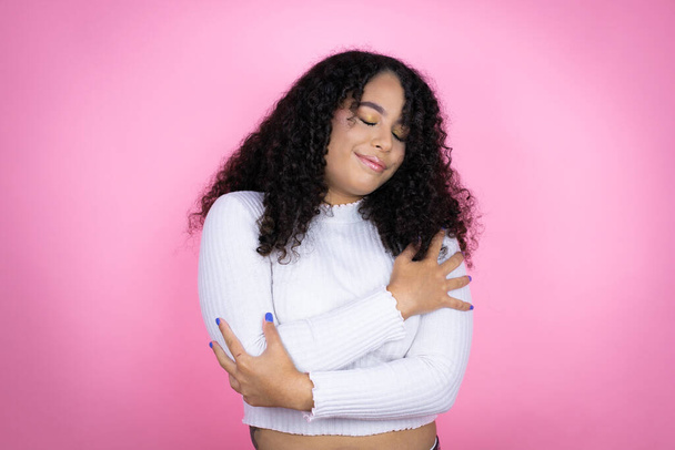 Afroamerikanerin trägt lässigen Pullover über rosa Hintergrund und umarmt sich glücklich und positiv, lächelt selbstbewusst - Foto, Bild