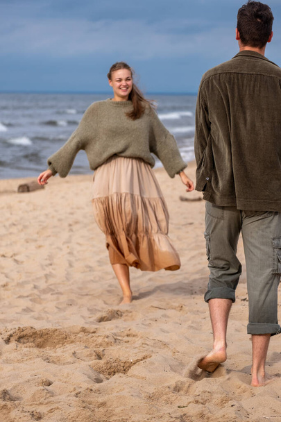 Couple strolling on beach, woman in beige skirt and green sweater, man in olive shirt. Mood of carefree leisure, suitable for travel or romance themes, wedding, engagement . High quality photo - Photo, Image