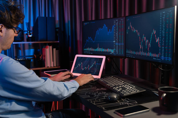 Joven inversor stock trader que busca en el ordenador portátil en comparación con las pantallas dinámicas de tipo de cambio del gráfico de mercado actual en línea en tiempo real en neón luz oficina moderna, el análisis de datos financieros. Gusher.. - Foto, Imagen