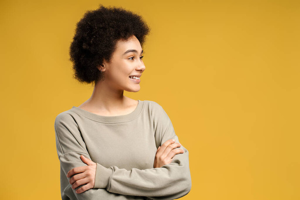 Kaunis, hymyilevä, nuori afroamerikkalainen nainen, jolla on kihara tukka ja kädet ristissä, katselee poispäin kopiotilasta, seisoo eristettynä keltaisella taustalla. Käsite ostoksia, mainos - Valokuva, kuva