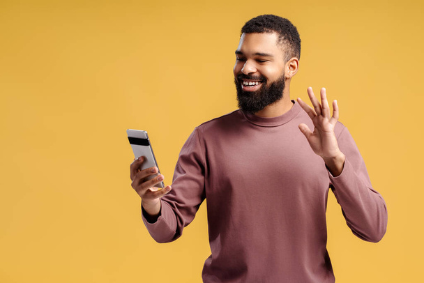 Jóképű, mosolygós, szakállas afro-amerikai férfi, blogger mobiltelefonnal, mobilalkalmazással integető kézzel, sárga háttérrel elszigetelt videófelvételekkel. Online technológiai koncepció - Fotó, kép