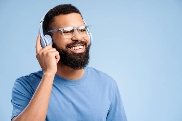 Attraente felice uomo afroamericano indossa occhiali alla moda ascoltando musica in cuffia guardando lontano copiare lo spazio isolato su sfondo blu. Concetto di pubblicità, hobby - Foto, immagini