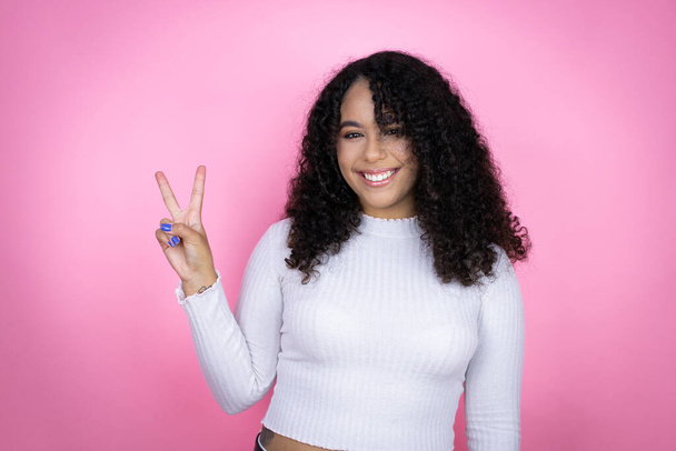 Afroamerikanerin trägt lässigen Pullover über rosa Hintergrund und zeigt mit Finger Nummer zwei nach oben, während sie selbstbewusst und glücklich lächelt - Foto, Bild