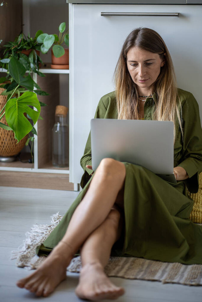 A relaxált szabadúszó nő, aki egy új projekten dolgozik a laptopon, a padlón ül, beltéri növényekkel körülvéve otthon. Koncentrált nő távirányítással a számítógépen. Diák nő néz oktatási webinárium. - Fotó, kép