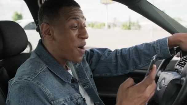 Heureux homme afro-américain lisant le message sur téléphone intelligent tout en conduisant la voiture. Personnes, mode de vie, concept technologique. Mouvement lent - Séquence, vidéo