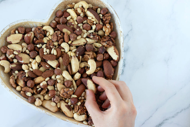 Hand holding Brazil nut and heart-shaped bowl full of mixed nuts isolated on white marble background. Top table view. Close-up. Healthy snacking, eating organic food concept. - Photo, Image