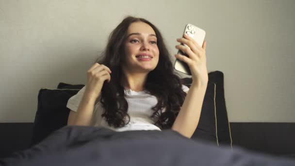 Onnellinen nainen pitää älypuhelinta puhuessaan verkossa ja katsoessaan ruudulle, istuu sängyllä. Hidasta vauhtia. Elämäntapa, teknologian käsite - Materiaali, video