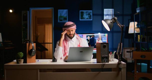 Zmęczony arabski biznesmen cierpiący na migrenę pracujący w biurze. Wyczerpany pracownik Bliskiego Wschodu w salonie odpowiadający na maile z przepracowaniem - Zdjęcie, obraz