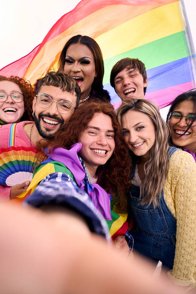 Svisle. Skupina mladých různorodých přátel se selfie při pohledu na přední fotoaparát mobilu slaví gay hrdost den společně drží duhovou vlajku. LGBT lidé komunita se usmívá a baví venku - Fotografie, Obrázek
