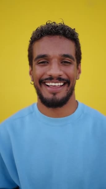 Verticaal videoportret van een jonge vrolijke Latijnse man met een stralende glimlach die met een vrolijke uitdrukking naar de camera kijkt, tegen een gele muurachtergrond. Mensen met positieve expressie - Video