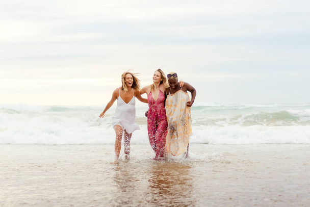 Багаторасова жіноча група з трьох друзів тримається разом, охолоджуючись у морській воді і насолоджуючись літнім днем на пляжі. Дружба, єдність і різноманітність - Фото, зображення