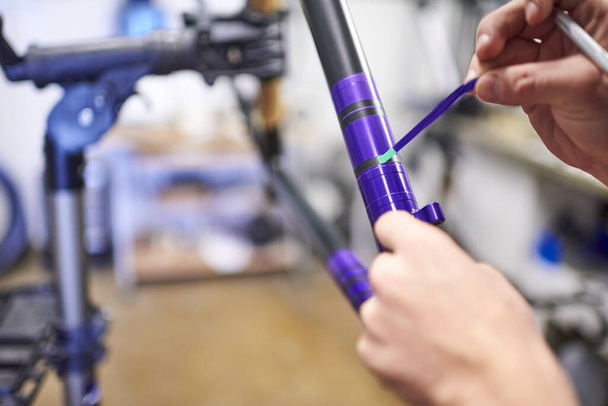 Persona desconocida que trabaja en un diseño de pintura de marco de bicicleta personalizado en púrpura y negro, un proceso creativo y técnico artesanal. Es hora de quitar las líneas de cinta adhesiva. Vista de cerca de las manos. - Foto, imagen