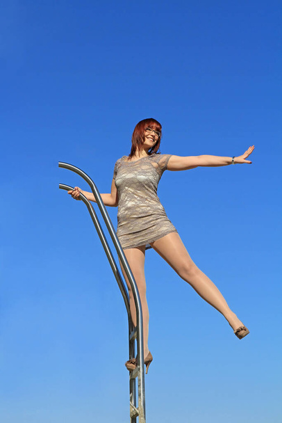 Una joven con un vestido se levanta en lo alto de una escalera y extiende un brazo y una pierna - Foto, Imagen