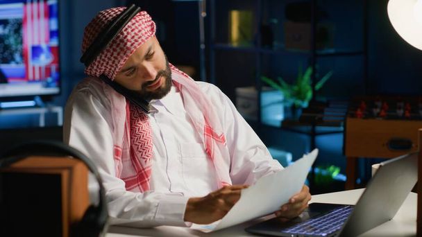 Arabski pracownik prowadzi rozmowy telefoniczne ze współpracownikiem podczas zdalnej pracy w przytulnym biurze. Telepracownik rozmawiający ze współpracownikiem za pomocą smartfona, omawiający papierkową robotę - Zdjęcie, obraz