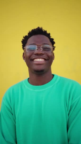 Verticaal videoportret van een jonge vrolijke Afrikaan met een stralende glimlach naar de camera kijkend met een vrolijke uitdrukking, tegen een gele muurachtergrond. Mensen met positieve expressie - Video