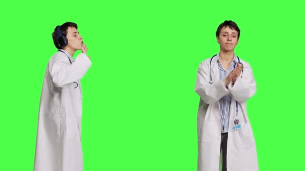 General practitioner clapping hands and listening to music on headset, standing against greenscreen backdrop. Woman specialist applauding someone and dancing around on cool songs. Camera B. - Footage, Video