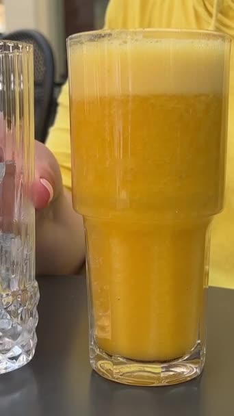 灰色のテーブルで氷の熱い日と美味しいオレンジジュース 女性手は,新しく絞った多品種の飲み物のスローモーションビデオ 小さな花のスポットに氷の断片をプールします. コルフ島で美しいサービング - 映像、動画