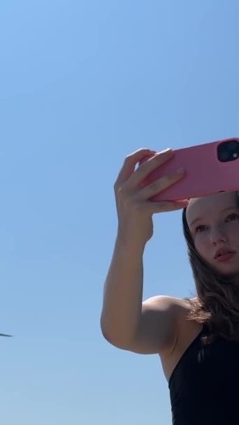 una joven adolescente. en una falda se para con un teléfono del iPhone XIII y dispara un vídeo de cómo un avión grande vuela cerca de su viaje Europa América adolescentes. - Imágenes, Vídeo