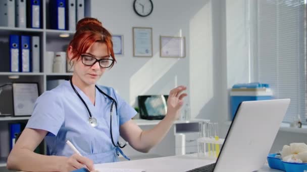 Ιατρικό εργαστήριο, νεαρή χαριτωμένη νοσοκόμα σε γυαλιά εξετάζει την αντίδραση του φαρμάκου σε δοκιμαστικούς σωλήνες και γράφει αναγνώσεις στο σημειωματάριο, ενώ κάθεται στο τραπέζι με φορητό υπολογιστή, χαμογελώντας και κοιτάζοντας κάμερα - Πλάνα, βίντεο
