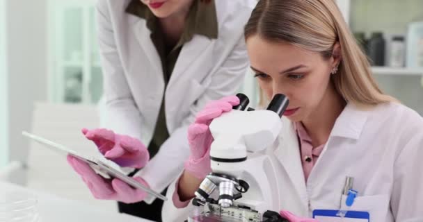 Lady kutató néz mikroszkóp és jóváhagyja a digitális jelentés tabletta kollégákkal a laborban. A tudósok a klinika technológiájával vizsgálják a mintákat - Felvétel, videó
